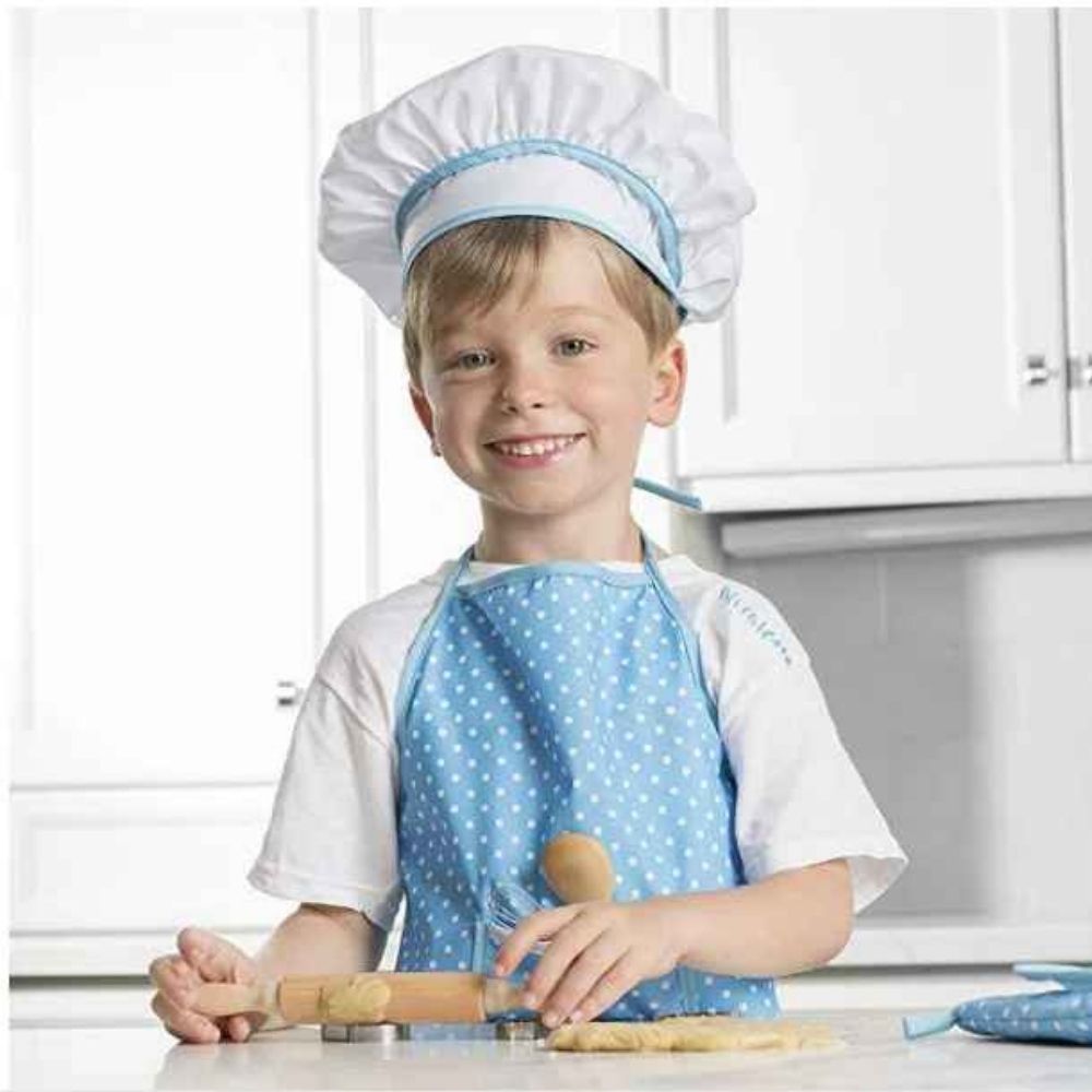 un garçon pourte son TABLIER CUISINE ENFANT de cuisinier bleu ainsi que son chapeau de grand chef pour jouer à la dinette enfant