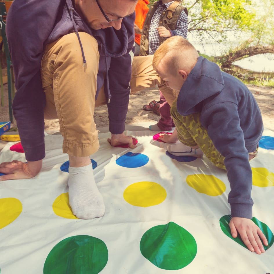 un père joue avec son fils au jeu de société Twister installé dans la cour